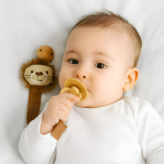 Un bébé couché sur un tissu blanc qui, mais ça susse dans sa bouche, accrochée par une attache tétine en forme de tête lion.
