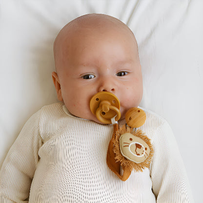 Un bébé couché avec une attache tétine en forme de tête de lion avec un chandail long blanc.