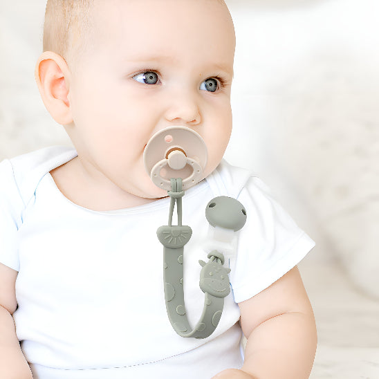 Un bébé avec une suce accroché par une attache tétine sur un chandail blanc devant un mur blanc