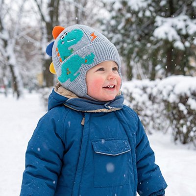 Un bébé portant un bonnet bebe bleu clair dehors en hiver avec de la neige qui tombe et des sapins.
