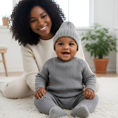 Un bébé portant un bonnet pour bebe au tricot gris, assis par terre avec sa maman.