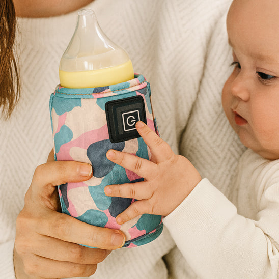 Un bébé dans les bras de sa maman avec un biberon dans un chauffe biberon portable dans la main.