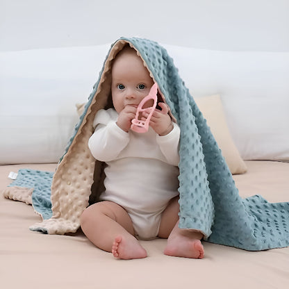 Un bébé assis sur un lit avec un jouet à la main avec une couverture sur la tête 