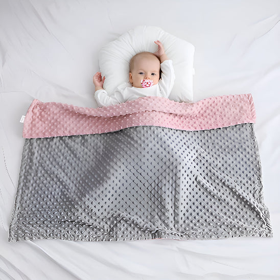 Un bébé avec une sucette dans la bouche, couché la tête sur un oreiller sur un lit avec une couverture gris et rose dessus lui 